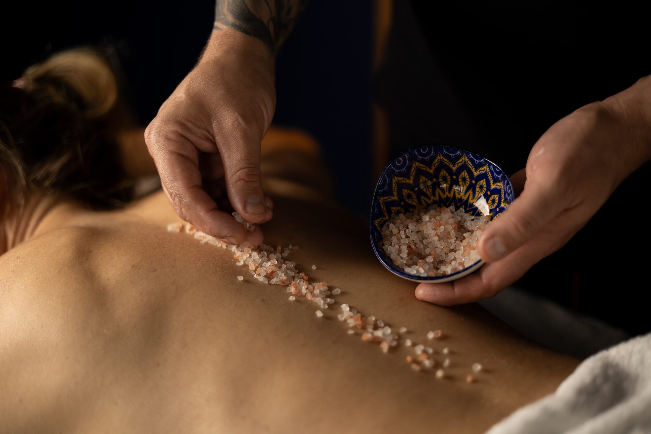 Terapeuta wykonujący masaż peelingujący na plecach klientki, używając naturalnych peelingów solnych w profesjonalnym gabinecie masażu